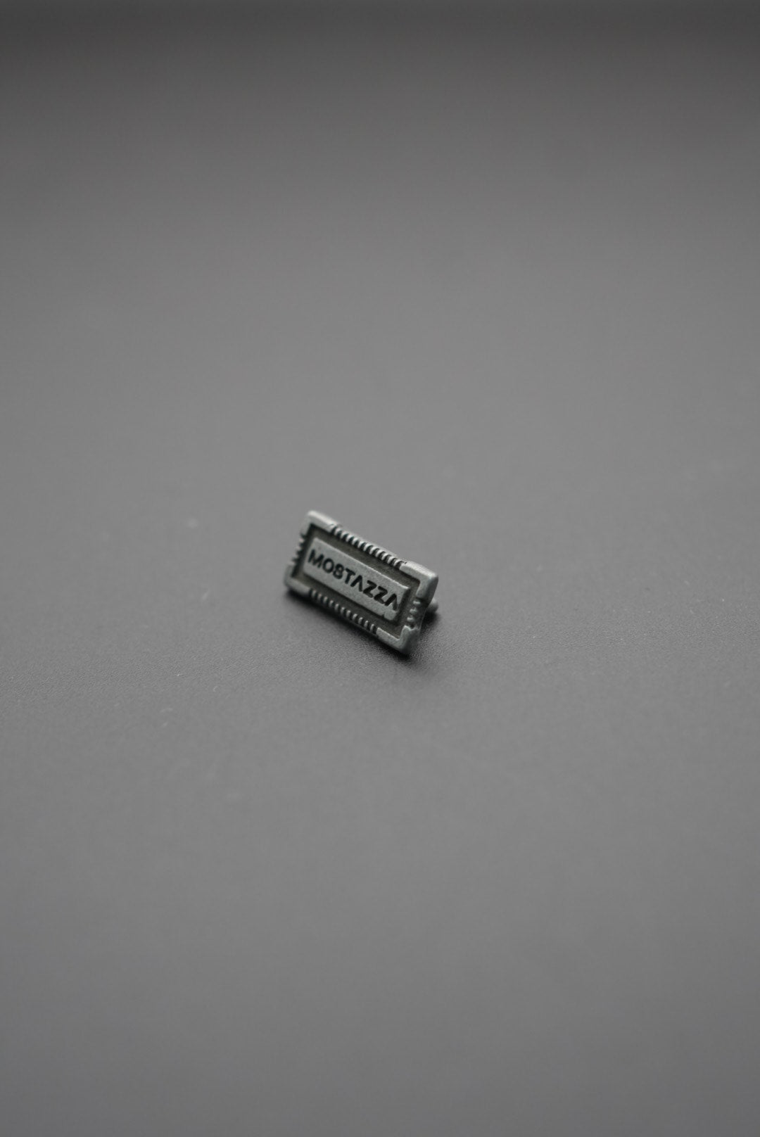 Etiqueta metálica rectangular con texto 'MOSTAZZA' en relieve sobre fondo gris