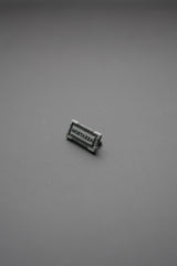 Vista frontal remache metálico rectangular con grabado MOSTAZZA sobre fondo gris