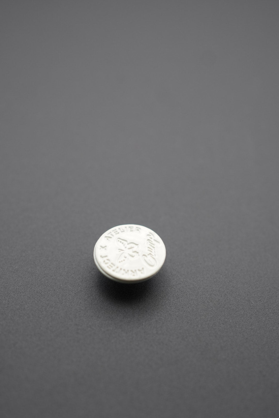 Detalle del botón Arkitect x Atelier con grabado claramente visible en fondo gris oscuro