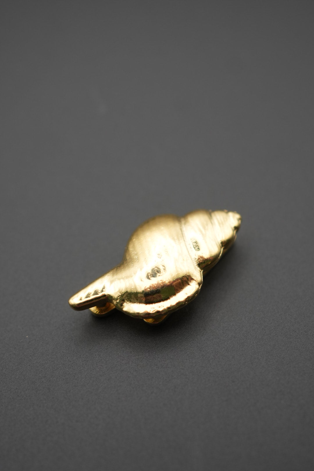 Pin metálico dorado en forma de concha marina sobre fondo gris oscuro vista frontal