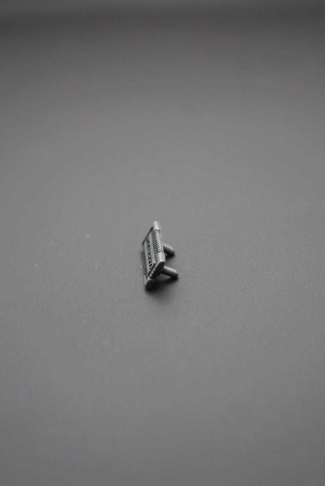 Vista lateral de etiqueta metálica rectangular con bordes texturizados y pin de sujeción