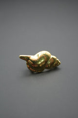 Pin metálico dorado en forma de concha marina sobre fondo gris oscuro vista frontal con enfoque en brillo