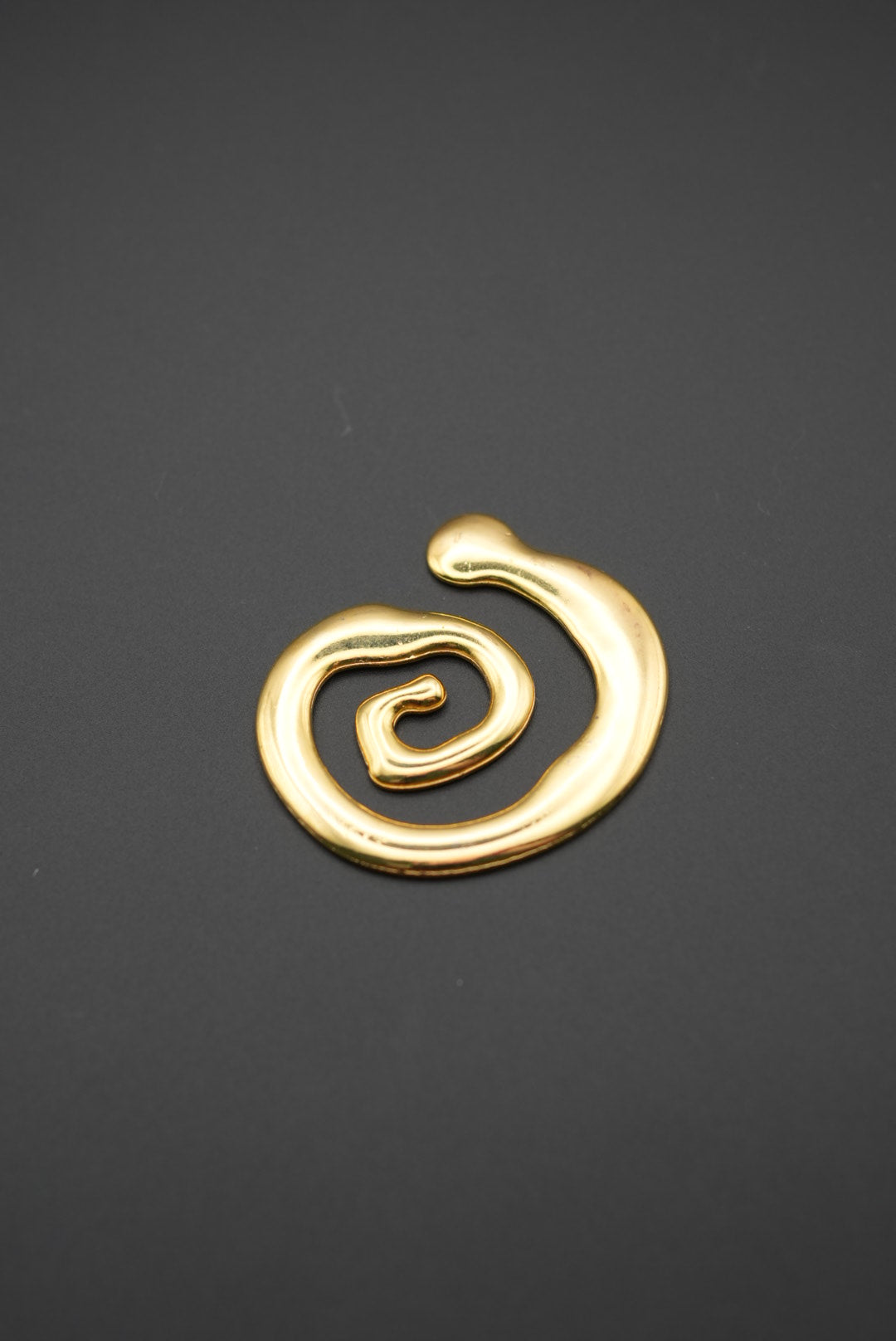 Vista detalle del broche dorado con diseño en espiral sobre superficie gris
