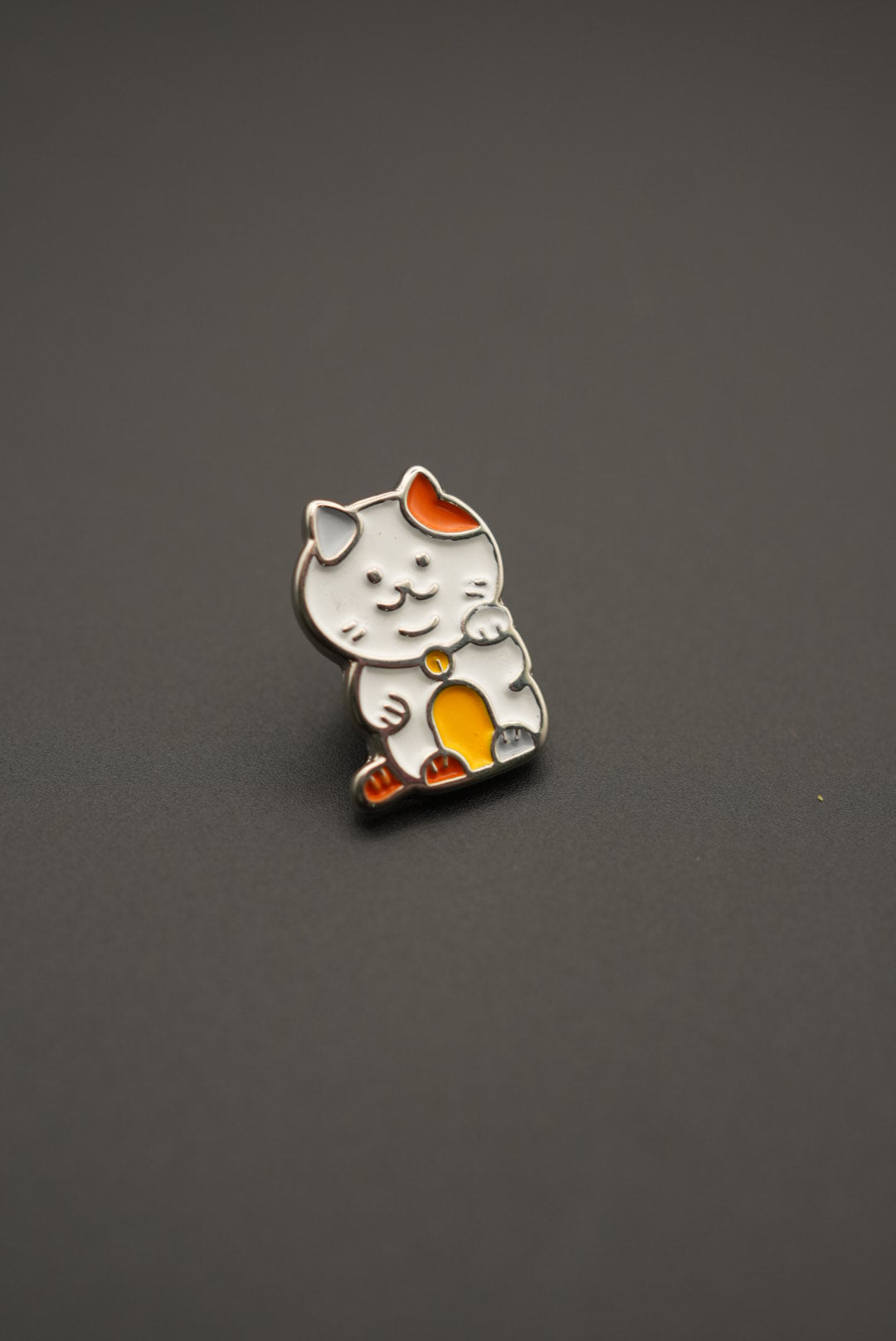Pin esmaltado gato Maneki-neko con detalles naranjas y amarillos presentado sobre fondo neutral
