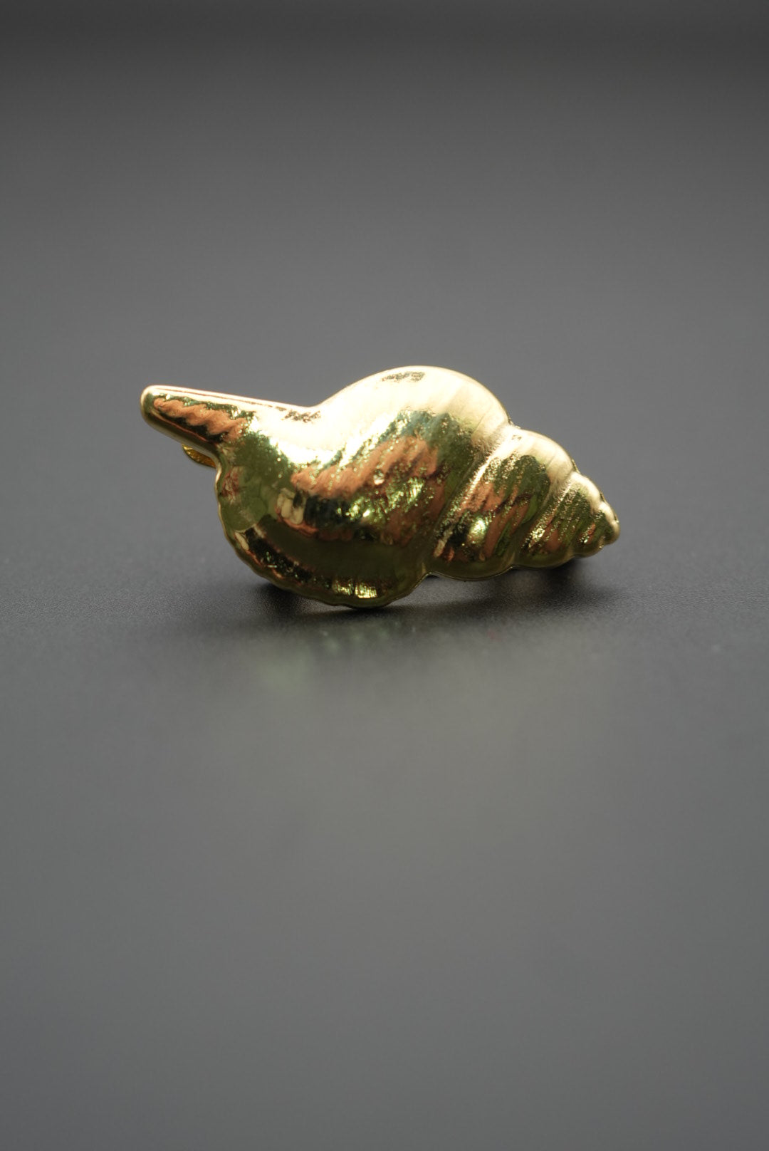 Pin metálico dorado en forma de concha marina sobre fondo gris oscuro vista lateral detallada