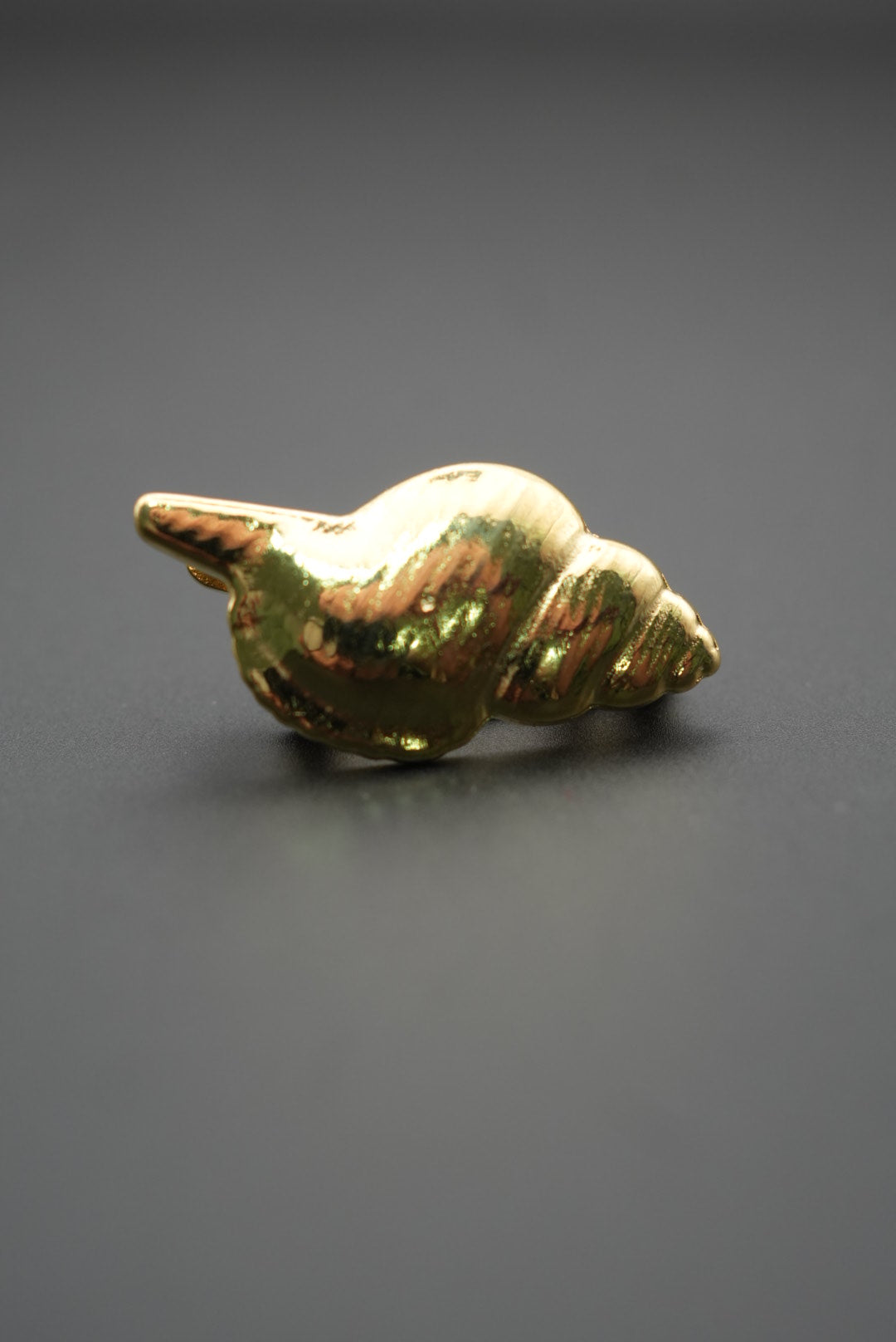 Pin metálico dorado en forma de concha marina sobre fondo gris oscuro vista lateral con reflejos