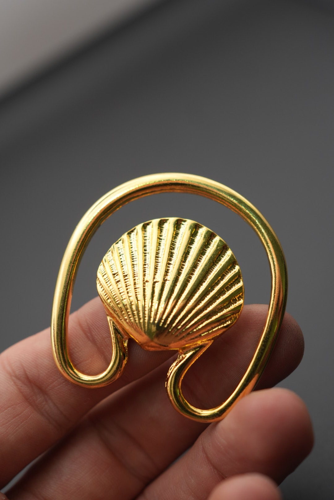 Gancho metálico de color dorado con diseño de concha detallada en mano, fondo negro