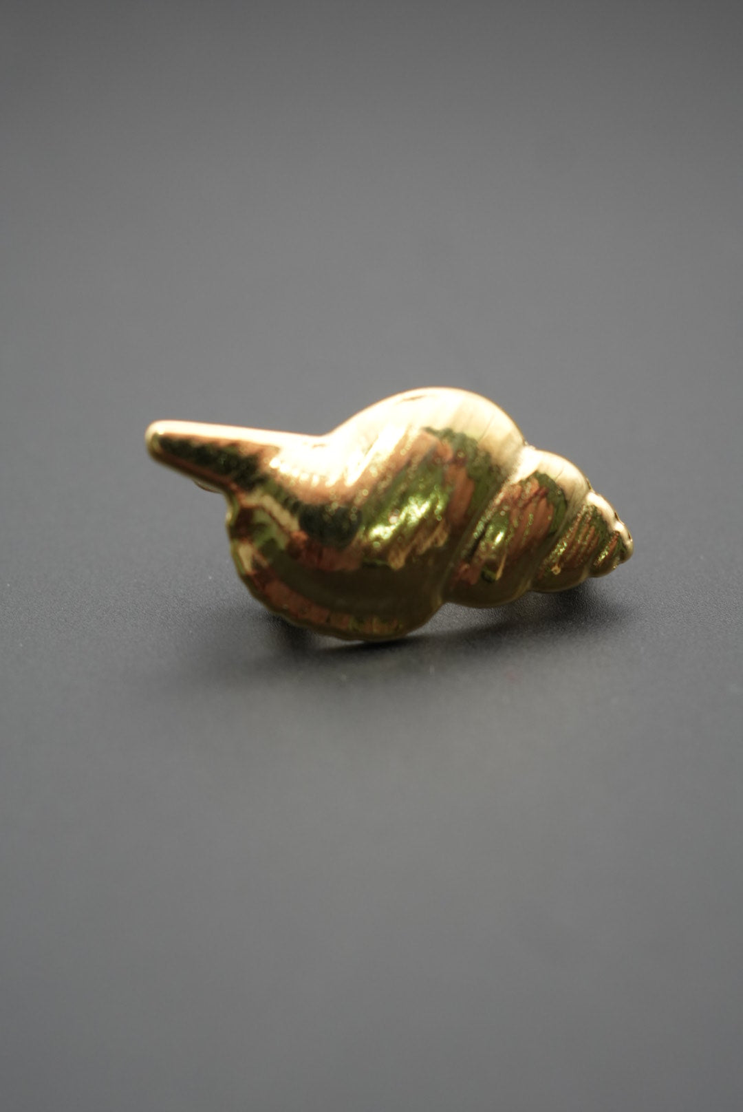 Pin metálico dorado en forma de concha marina sobre fondo gris oscuro vista lateral inversa